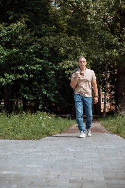 Vertical stylish relaxed, confident mature gray haired businessman in sunglasses with jacket on shoulder in casual clothes walking street near park. Fashion street style male mode, weekend activity