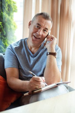 Vertical smiling positive grizzled man using mobile phone and make notes in diary textbook, look camera in cafe. Online remote business, trader and sales occupation. E-learning and distance education