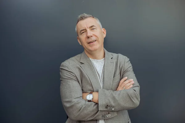 Close up smiling, confident mature gray haired man with crossed arms look at camera, presenting new product, service against gray wall. Innovative ideas. Promotion, victory, great idea. Copy space