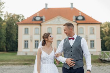 Charming, blissful, joyful married couple of woman in glasses and white lace wedding dress and veil with man in suit on wedding celebration against house. Moving house, mortgage and for young family