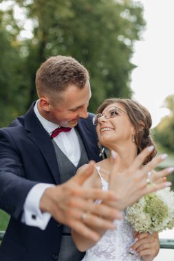 Vertical glad, fortunate, blissful married couple on wedding celebration showing and brag about wedding rings on hands on foreground. Newlywed, new traditional family. Celebration in natural parkland