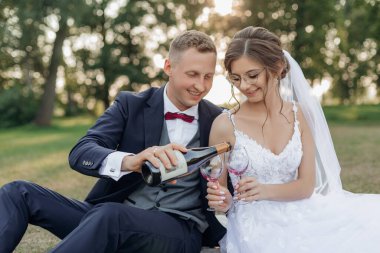 Smiling and shy groom and bride drinking and pouring bubbly champagne wine from bottle to glasses together, sitting on blanket in park and having fun on wedding celebration day. New family goals