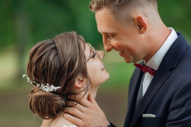 Close up affectionate, cheerful, smiling bride and groom caring each other, embracing, standing face to face on wedding celebration on green background in park. Bounding relationship, love, romance