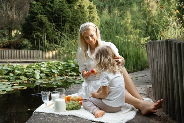 Kind, attractive woman feeding little girl on picnic, having organic fresh fruit and milk lunch in natural park near lake. Healthy organic diet dairy food. Health care and active lifestyle for family