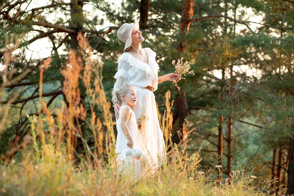 Charming, gentle woman, mother in hat and little blonde girl in dresses walking together in sunny natural pine forest. Ecological vacation and holiday with family, healthy and active lifestyle