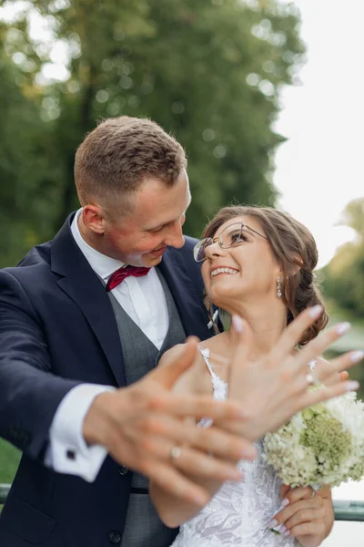 Vertical glad, fortunate, blissful married couple on wedding celebration showing and brag about wedding rings on hands on foreground. Newlywed, new traditional family. Celebration in natural parkland