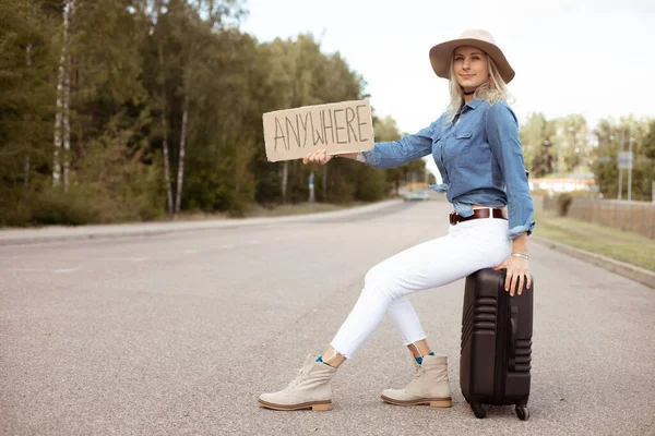 Nice woman try to stop passing car sitting on suitcase with outstretched cardboard poster on empty highway. Lady in hat escape from city to go anywhere. Travelling, hitchhiking, vacations, autumn.