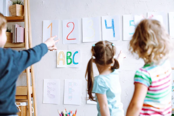 Children stand with back and learn english letters, alphabet and numbers on cards on wall. Kids point at colored numbers on sheets of paper by finger. Reading and counting, study in daycare centre.