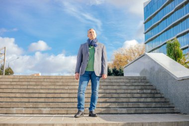 Old handsome businessman in stylish scarf stand on paved stairs on blue sky background, from below view. Man walk outdoors. Urban lifestyle, modern infrastructure, autumn season, leisure.