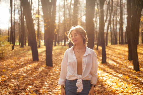 Cheerful young woman in golden forest among bare trees and yellow fallen leaves. Person enjoy leaf fall walking in autumn park on sunny day. Beautiful fall nature, bright sunlight, lifestyle.