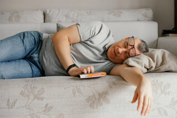 Aged man with glasses fall asleep reading book on comfortable couch in home room. Daytime sleep, napping. Tired old person lying head on pillow on sofa. Stylish soft furniture, relaxation.