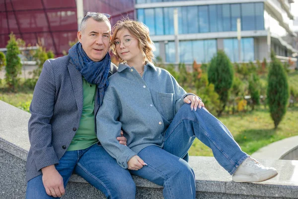 Portrait of adult 50 years old man sitting on bench with teenage girl on street near green plants. Father and daughter relax, family walking. Urban lifestyle, outdoor recreation, leisure.