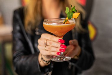 Woman with red manicure in black jacket hold glass of fresh alcoholic cocktail on blurred background closeup, selective focus. Cold sweet drink with mint and orange decor. Holiday, relax, bar.