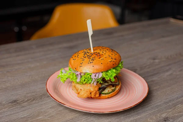 Closeup plate with juicy appetizing chicken burger isolated on wooden table background. Serving of big caloric sandwich on skewer. Fast food cafe, unhealthy nutrition, restaurant dish, dinner.