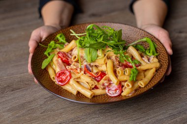Hands of human hold plate of delicious italian pasta with arugula and tomatoes in delicate creamy sauce on table background closeup. Serving and decor of restaurant dish. Gourmet european food.