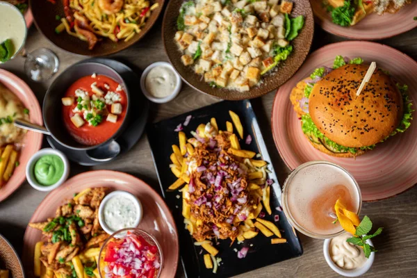 Table full of delicious food in plates top view. Fries, soup, salad, burger, sauces and drinks on festive dinner for event celebration. Nourishing food, menu of restaurant, beautiful serving food.