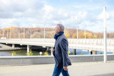 Calm gray haired man looking away camera in warm jacket and scarf walking street, near promenade bridge. New winter, autumn season and low temperature, cold and flu, sick and illness in the city. 