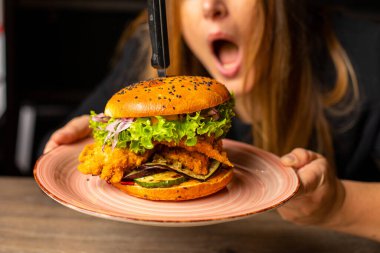 Cropped photo of shocked woman with open mouth holding big pink plate with tasty burger with lettuce, red onion, tomato, fried chicken punctured with knife. Cooking, fast food, junk food, emotion.