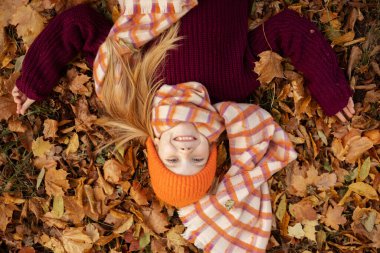 Glad, satisfied, delightful little blond girl in warm outfit, scarf and hat lying and play with colorful leaves on autumn foliage. Fashionable warm wool clothes for children. Top view, high angle view
