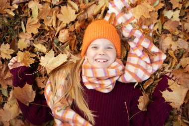 Positive, glad, toothy smiling, delightful little blond girl in warm outfit lying and holding colorful leaves on autumn foliage. Trendy and warm wool clothes for children. Top view, directly above
