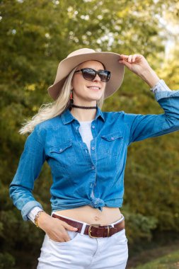 Vertical smiling, modern, confident, glad, carefree blond woman in sunglasses and hat looking at sun and joying walking and fresh nature air on vacation holiday. Relaxed traveler and tourist activity
