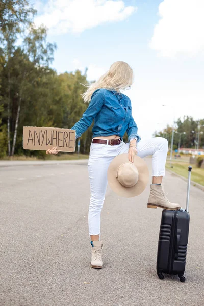 Woman with suitcase and cardboard poster stop passing car on empty highway and look back. Lady in hat escape from city to go anywhere. Travelling, free transportation, hitchhiking, vacations.