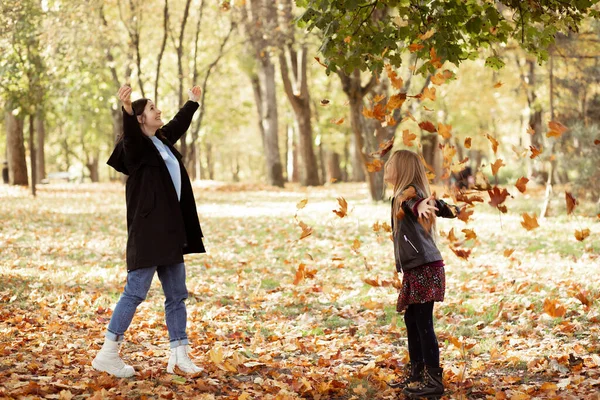 Playful, pleased, funny, glad, carefree mother and daughter playing with leaves, throw like rain in golden autumn woodland. Family of two having fun before school in cold season. Weekend activity