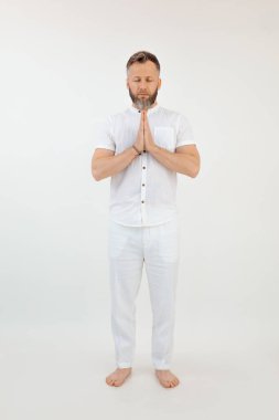 Portrait of concentrated sporty barefoot middle-aged bearded man with short grey dark hair, closed eyes wearing white shirt, trousers, standing, folding hands on white background. Meditation. Studio.