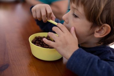 Pretty little boy sitting at brown wooden table in kitchen and having breakfast closeup. Kid of kindergarten age holding spoon to eat chocolate balls with milk. Ready breakfast, health nutrition.