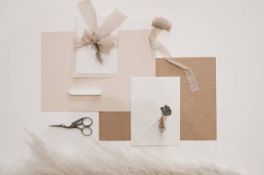 Top view of composition of white, brown, pink sheets of paper, scissors, white envelope with grey wax seal, bow of transparent ribbon, pink skein of ribbon and dried pampas grass on white background. 