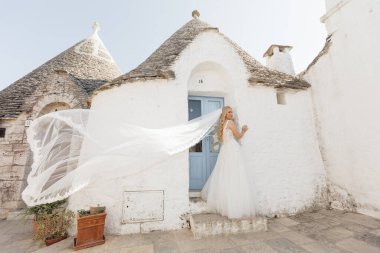 Side view of young woman bride with long fair hair wearing wedding dress, long flying gorgeous bridal veil, standing near blue door of old white stone house trullo with conical roof in Italy. Wedding.