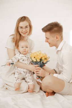 Handsome father gift little baby and mother bouquet of yellow tulips sitting on bed on white background. Home family photo of mom, dad and infant child. Family time, holiday, parental love.