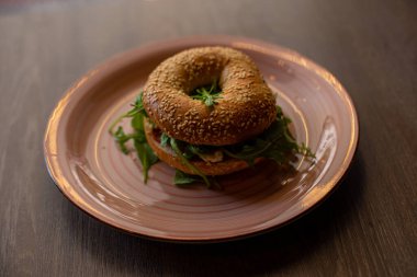 Close-up of pink ceramic plate with fresh palatable burger with hole in bun, meat, arugula on wooden table. Cooking, restaurant, menu, advertising, fast food, junk food, cafe, delivery, promotional.