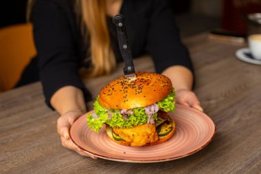 Cropped photo of woman sitting at table, holding plate with yummy burger with lettuce, red onion, cucumbers, fried chicken punctured with knife. Cooking, restaurant, menu, fast food. Selective focus.