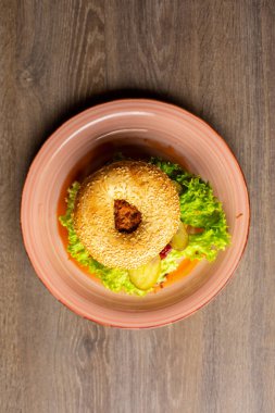 Top view of pink ceramic plate with fresh appetizing burger with hole in bun, lettuce, fried chicken, pickled cucumbers on wooden table. Cooking, restaurant, menu, advertising, fast food, junk food.