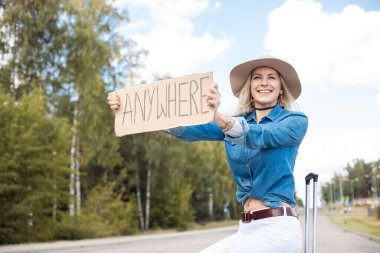 Positive, smiling, funny, carefree blond woman in cowgirl hat and jeans shirt hitchhiking, attract attention by road with cardboard plate anywhere sit on baggage suitcase and wait car share traffic