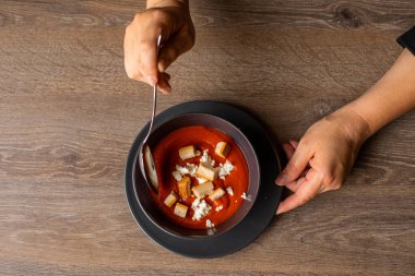Top view of unrecognizable woman holding and melting savory tomato soup with sour cream and croutons in black bowl with spoon on brown wooden table. Cooking, vegetarian soup, advertising, delivery.