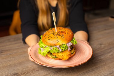 Cropped photo of woman sitting at table, holding big pink plate with fresh palatable burger with lettuce, red onion, tomato, chicken. Cooking, restaurant, menu, advertising, fast food. Soft focus.