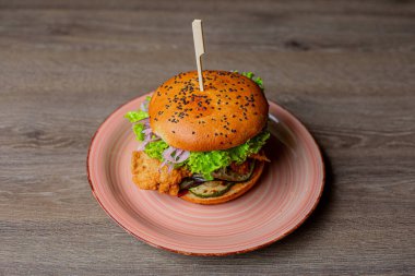 Close-up of pink ceramic plate with fresh toothsome burger with lettuce, red onion, tomato, chicken punctured with stick on wooden table. Cooking, restaurant, menu, advertising, fast food, junk food.