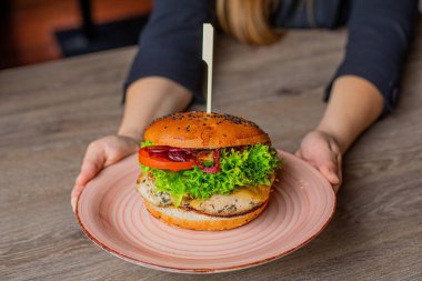 Cropped photo of woman sitting at table, holding big pink plate with fresh delicious burger with lettuce, red onion, tomato, chicken. Cooking, restaurant, menu, advertising, fast food, junk food.