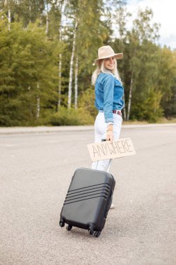 Vertical smiling, cheerful blond woman hitchhiker with carton plate anywhere carrying roll aboard suitcase and waiting car stop, walk by asphalt road. Adventure hitchhiking lifestyle. Travelling time