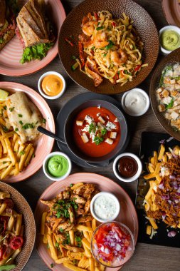 Top view of colorful food on wooden table in restaurant cafe. Pasta with shrimps. Caesar salad with chicken. Sandwiches. Various sauces. Tomato soup. French fries, omelette, grilled meat. Vertical.