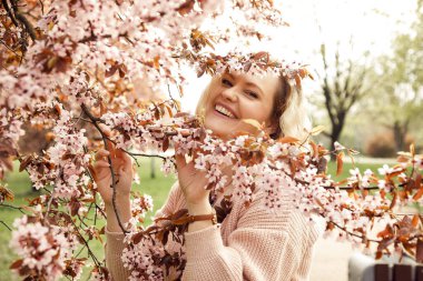 Portrait of young joyful laughing attractive woman with long wavy fair hair wearing pink knitted cardigan, standing near blossoming bird-cherry tree, holding branch in park, having fun. Spring. 
