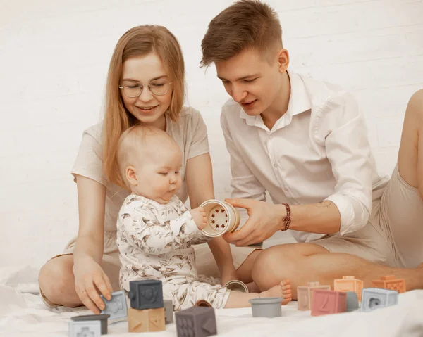 Happy family of three people sit on bed and play toys with little baby on white background. Home family photo of mom, dad and infant child. Concept of parental affection and caring for children.
