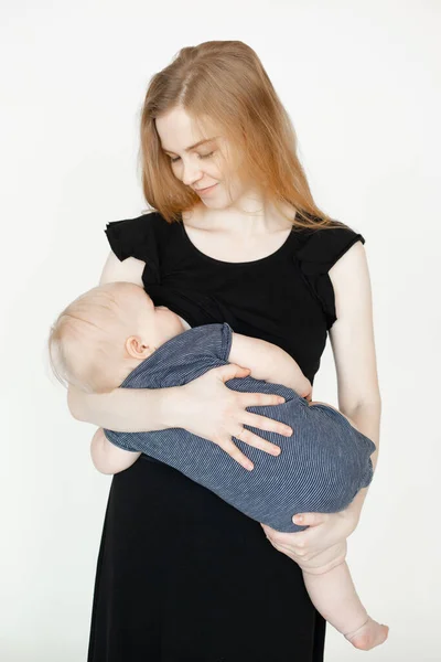Young mother in black dress breastfeed infant baby and with smile look down, white background. Portrait of lying on hands little child sucking breast milk. Maternal love, lactation, breastfeeding.