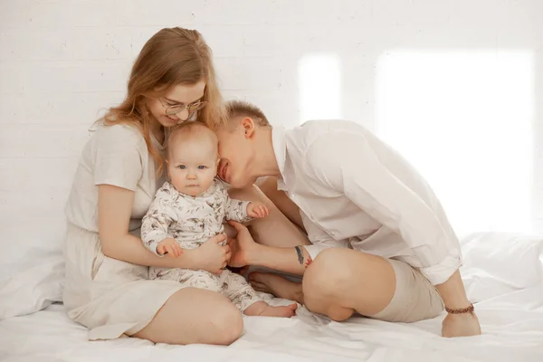 Portrait of happy family of three on white background. Mother embrace and father with love kiss infant baby. Photo of young couple and child in white family look. Parental affection, childhood.
