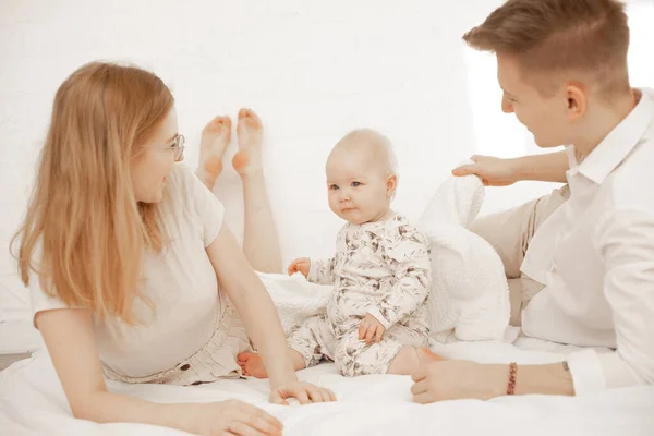 Portrait of happy family of three on white background. Mother, father and infant baby lie on bed. Photo of young couple and child in white family look. Parental affection, childhood, love.