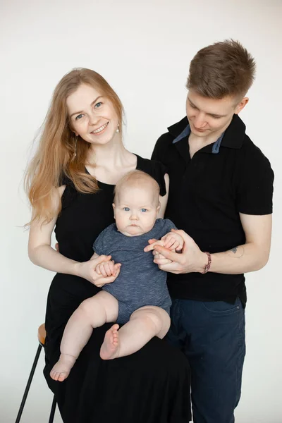Happy family from three in black looks on white background. Mom with beautiful smile sit on chair and hold infant baby, dad stand near. Family photo, parenting, firstborn, love, feelings.