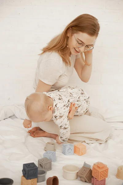 Smiling young mother sit on laps on bed and hold little baby with tummy down on white background. Home family photo of playing woman and infant child. Concept of maternal affection and childcare.