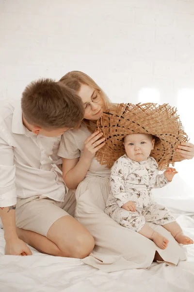 Young family couple sit on bed and dress up little baby wicker hat on white background. Home family photo of mom, dad and infant child. Family time and concept of parental affection and love.